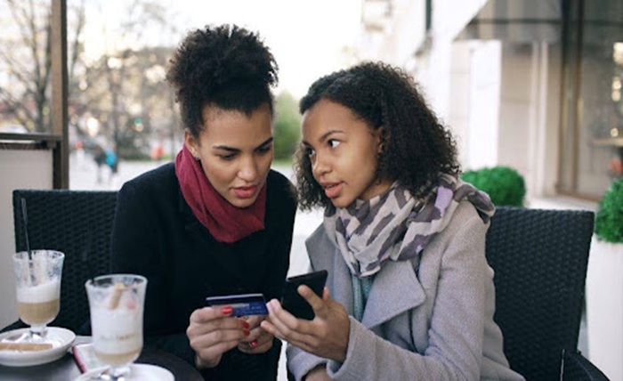 two women shopping online with a smartphone and credit card at an outdoor cafe
